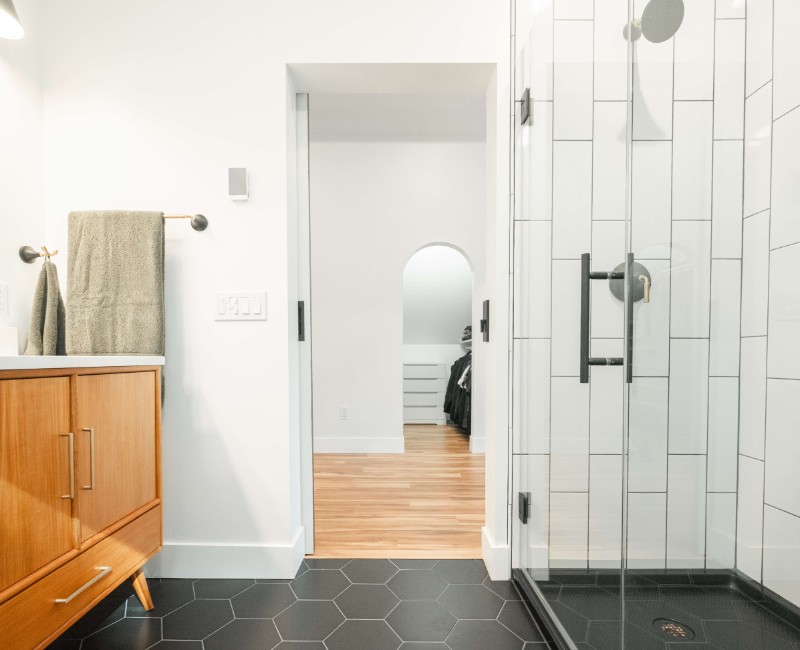 A bold black-and-white ensuite bath with frameless glass, warm wood cabinetry, and a custom sliding door — part of a NARI award-winning attic conversion by Chads Design Build in Madison, WI.