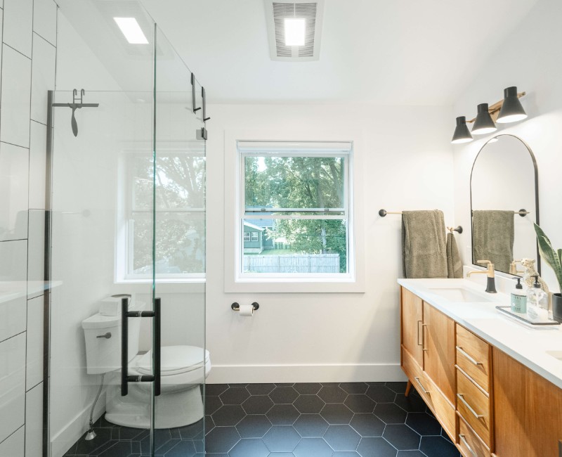 A bold black-and-white ensuite bath with frameless glass, warm wood cabinetry, and a custom sliding door — part of a NARI award-winning attic conversion by Chads Design Build in Madison, WI.