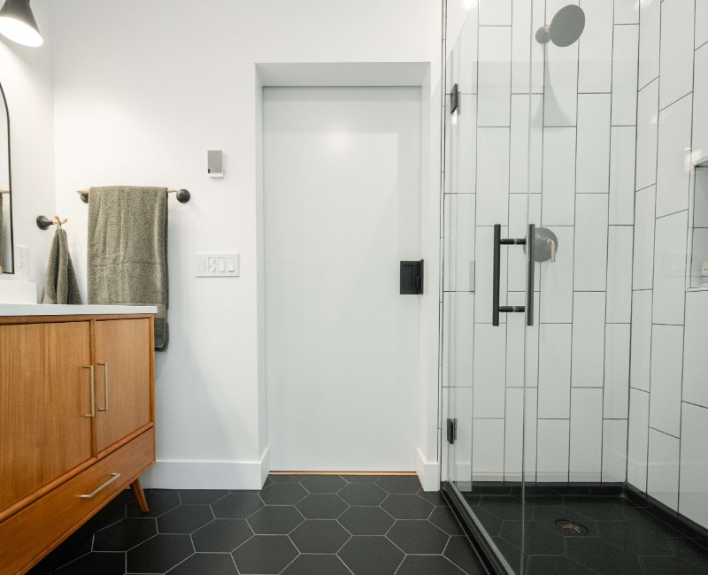 A bold black-and-white ensuite bath with frameless glass, warm wood cabinetry, and a custom sliding door — part of a NARI award-winning attic conversion by Chads Design Build in Madison, WI.